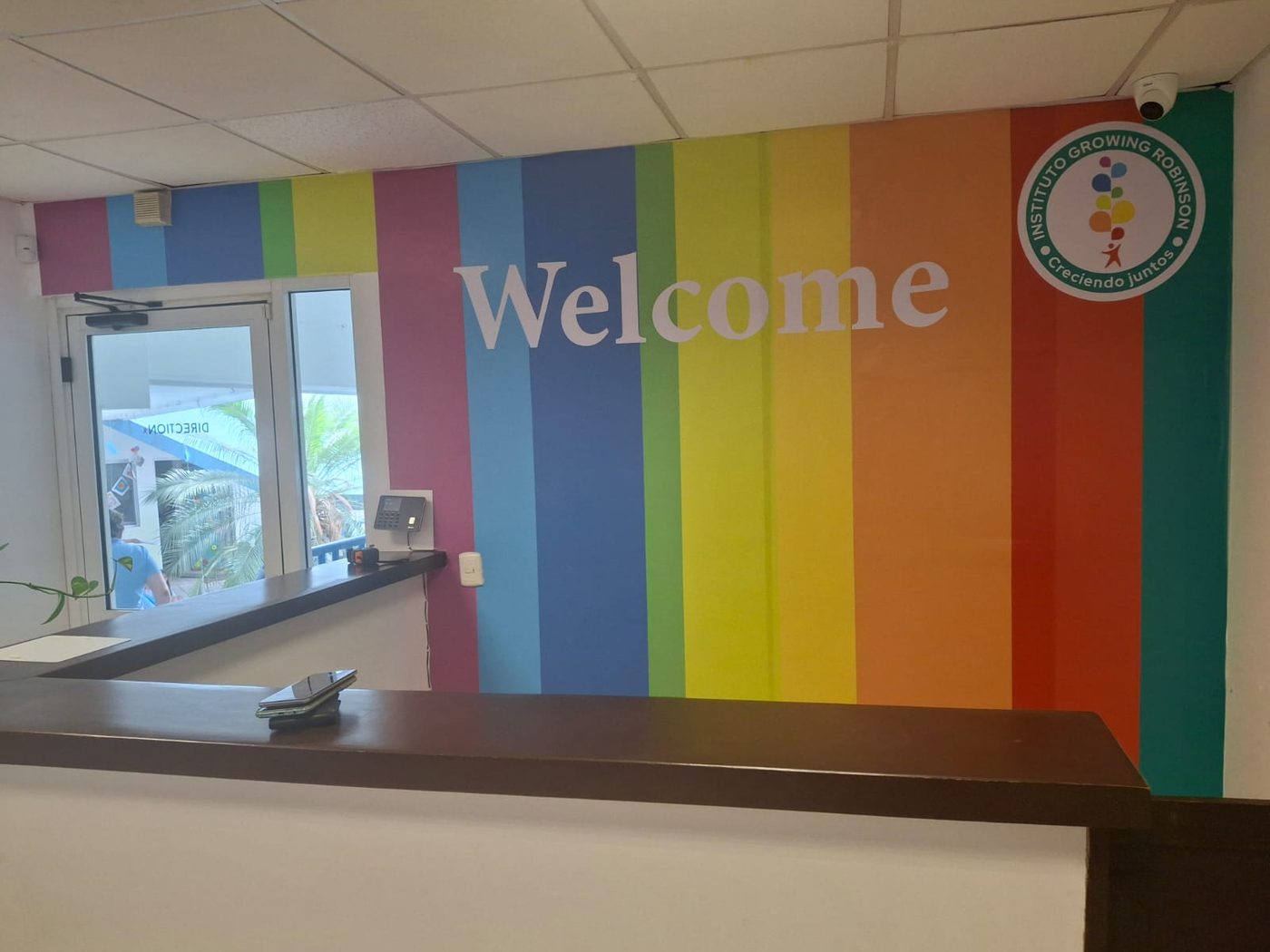 Mural de recepción del Instituto Growing Robinson con tipografía 'Welcome' grande en blanco sobre franjas verticales de colores arcoíris instalado por Punto Clave