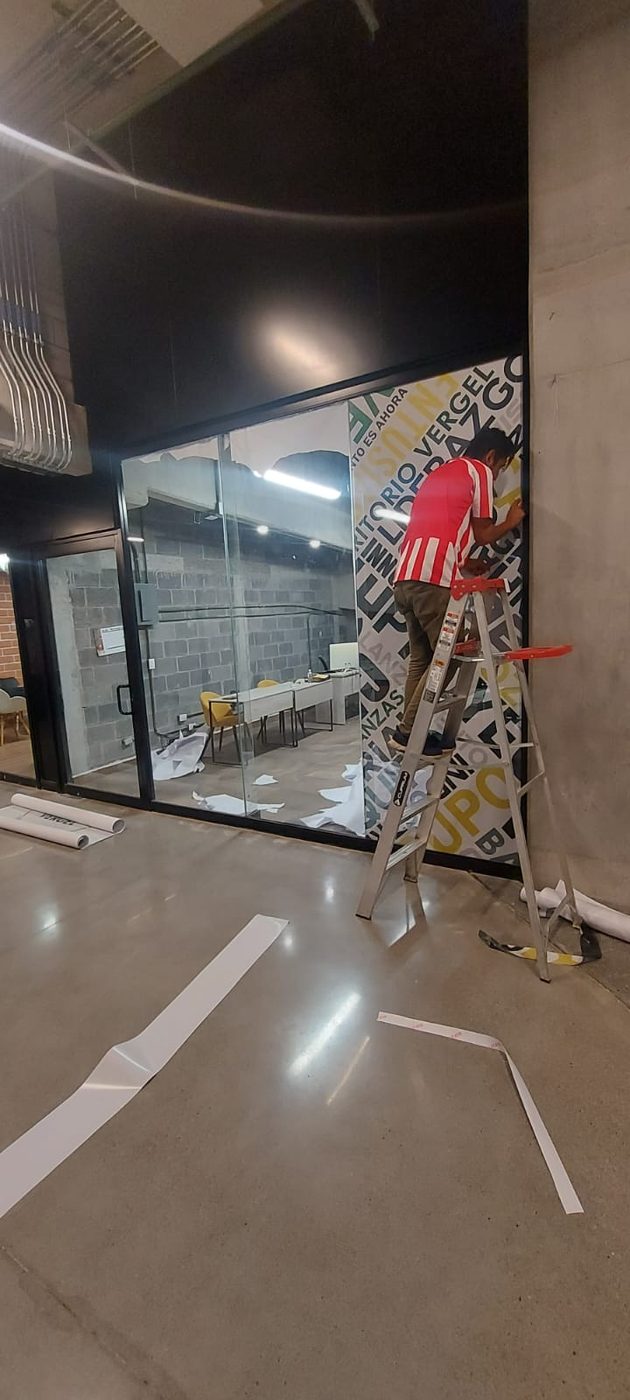 Técnico de Punto Clave instalando vinil tipográfico de gran formato sobre cristal corporativo de oficinas durante proceso profesional con escalera y herramientas en Monterrey